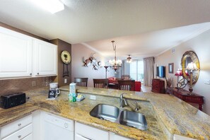 A kitchen with granite countertops, a double sink, and white cabinetry. There is a coffee maker, a toaster, and a dish rack on the counter. - Coastal condo steps to beach with pools, hot tub, gym & amazing views (Perdido Key)