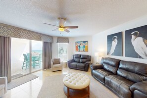A living room with a ceiling fan, a leather sofa, a tufted ottoman, and a dining area with a table and chairs. - Coastal condo with beach, pool, hot tub, sauna, tennis, bikes, W&D, & central AC (Santa Rosa Beach)