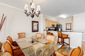 A dining area with a glass table and chairs, a chandelier, and a kitchen in the background. - Condo with 2 stunning pools, hot tub, gym, partial Gulf view & pickleball (Santa Rosa Beach)
