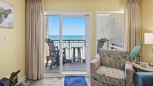 Une pièce comprenant un canapé, une petite table, une chaise, un tapis bleu et une vue sur l’océan, visible à travers une porte coulissante.
