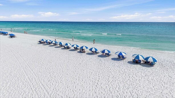 Une plage bordée de rangées de parasols bleus et blancs, avec du sable blanc et l’océan.