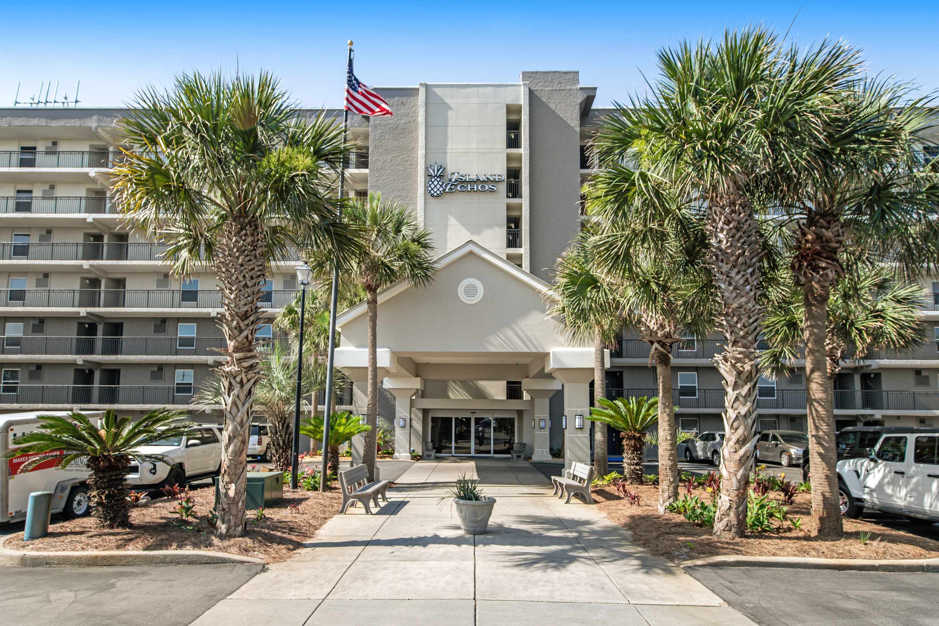 A hotel entrance with a flag, palm trees, and a parking area.