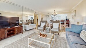 Un vaste salon doté d’un téléviseur, d’un canapé et de fauteuils, qui donne sur un espace cuisine.