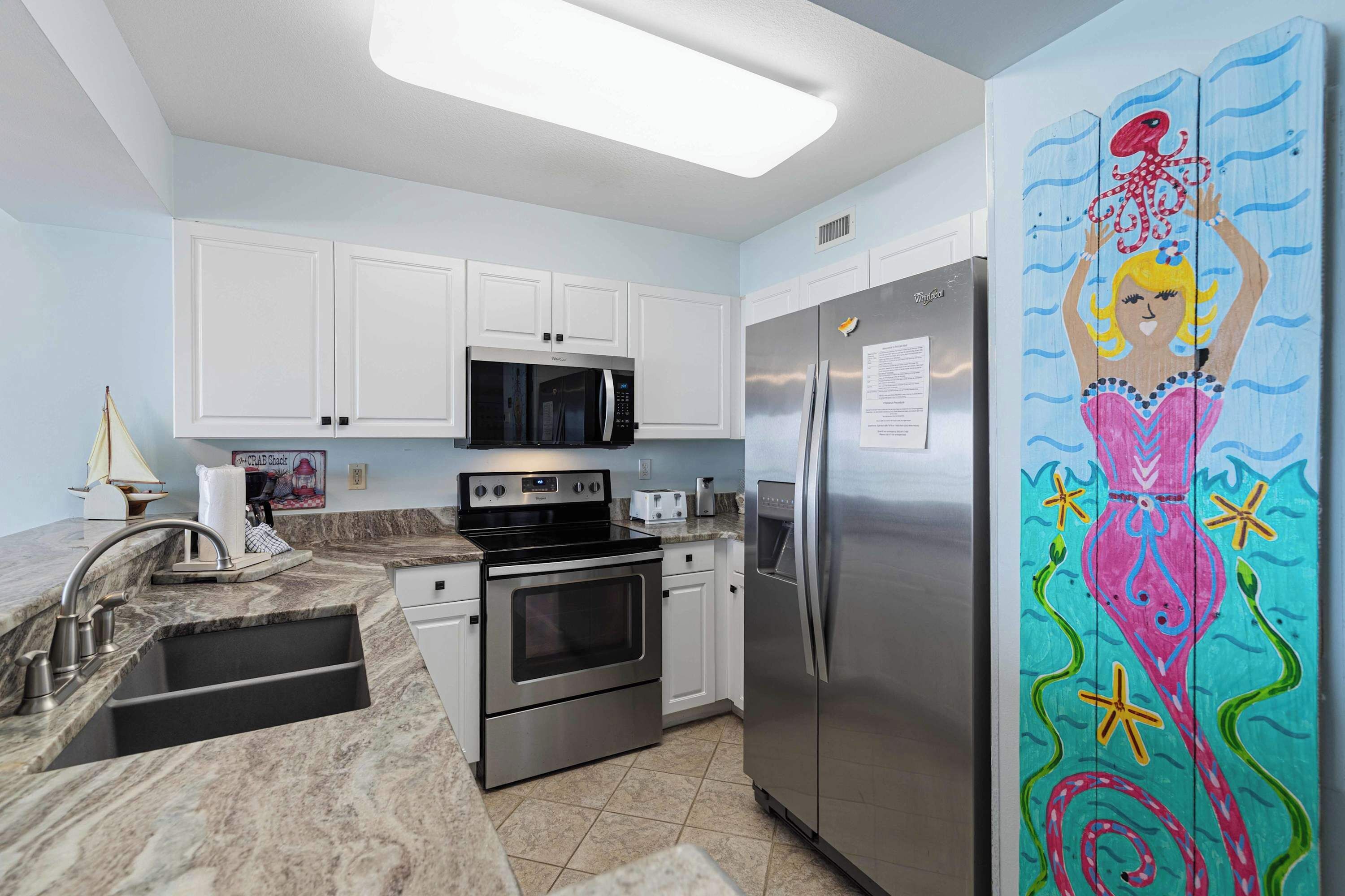A kitchen with white cabinets, stainless steel appliances, a marble countertop, and a colorful mermaid mural on the refrigerator.