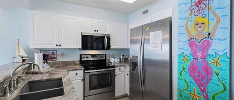 A kitchen with white cabinets, stainless steel appliances, a marble countertop, and a colorful mermaid mural on the refrigerator.