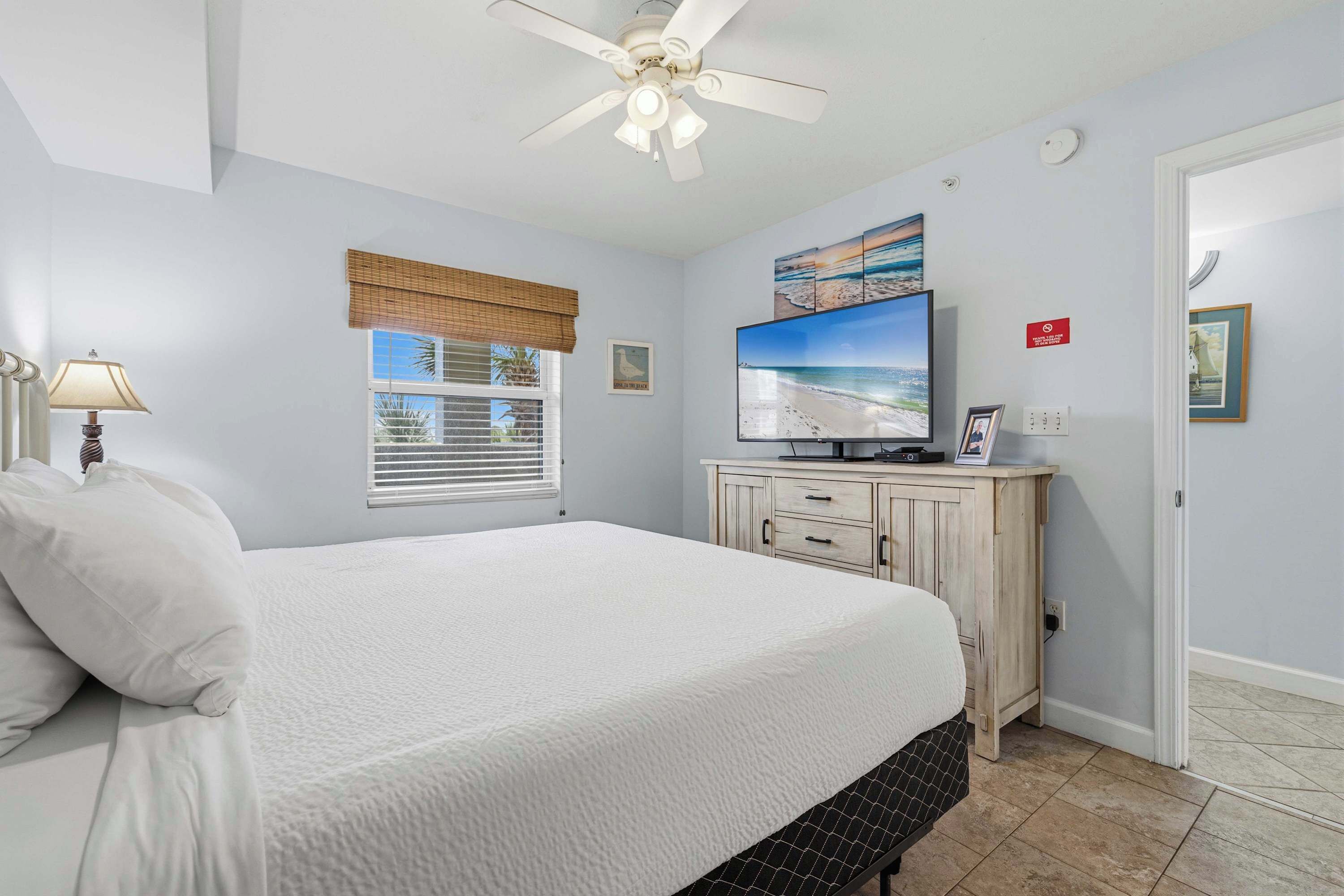 A bedroom with a bed, a ceiling fan, a television, a window with blinds, and a door.