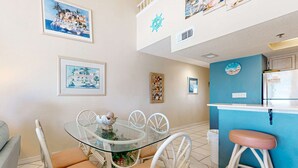 A dining area with a glass table and white chairs, a kitchen with a blue accent wall, and artwork on the walls. - Gulf-front condo with complimentary beach equipment - near shops & dining (Destin)