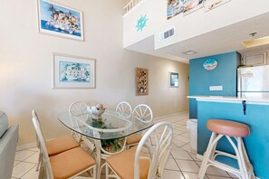 A dining area with a glass table and white chairs, a kitchen with a blue accent wall, and artwork on the walls.