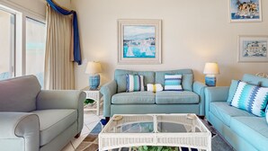 A living room with a light blue sofa, two side tables with lamps, a glass coffee table, and a view of the sea through large windows. - Gulf-front condo with complimentary beach equipment - near shops & dining (Destin)