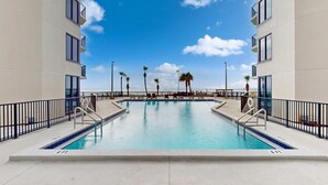 A swimming pool with a clear view of the ocean, surrounded by a building with balconies and palm trees. - Gulf-view condo with furnished balcony, pools, beach access, fitness room (Panama City Beach)