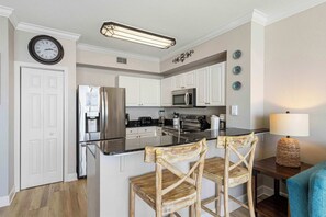 A modern kitchen with white cabinets, stainless steel appliances, and a central island with two wooden chairs. - Beachfront Condo, gorgeous views, shared pool and beach access (Panama City Beach)