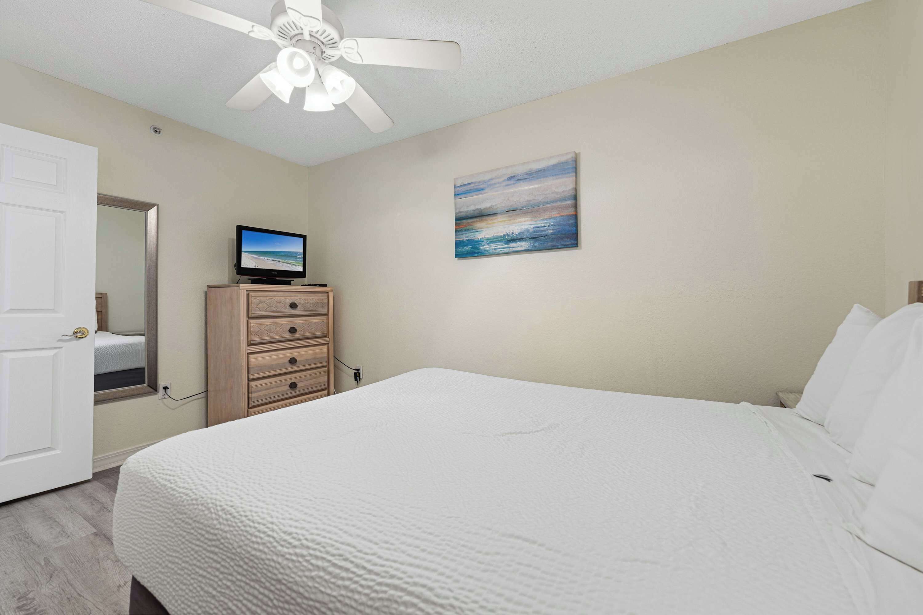Une chambre à coucher avec un lit, un téléviseur posé sur une commode en bois, un ventilateur de plafond et un tableau accroché au mur.