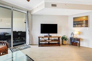A modern living room with a flat-screen TV on a wooden stand, a glass coffee table, a sofa, a dining area with chairs, and a painting of a sunset on the wall. - Oceanfront condo with pool, hot tub, sauna, gym, bikes, tennis - lovely balcony (Santa Rosa Beach)