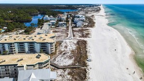 Luftaufnahme einer Küstenlandschaft mit Strand, einer Straße, die zu einer Gruppe von Gebäuden führt, und einem Gewässer im Hintergrund.