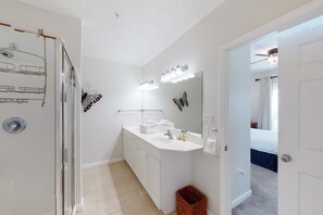 Een moderne badkamer met een wit wastafelmeubel, een grote spiegel met verlichting, een douchegedeelte en een zichtbare deur naar een andere kamer.