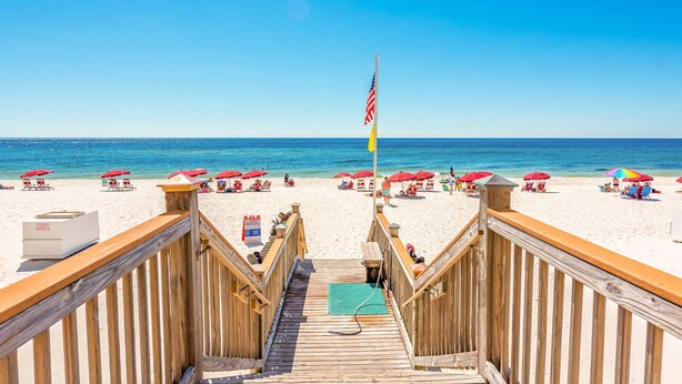 Ein Holzsteg führt zu einem Strand mit Sonnenschirmen und einer Flagge.
