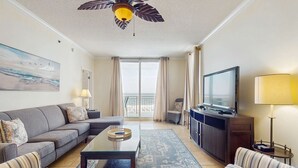 Een woonkamer met een bank, fauteuils, een salontafel, een televisie, een plafondventilator en een balkon met gordijnen.
