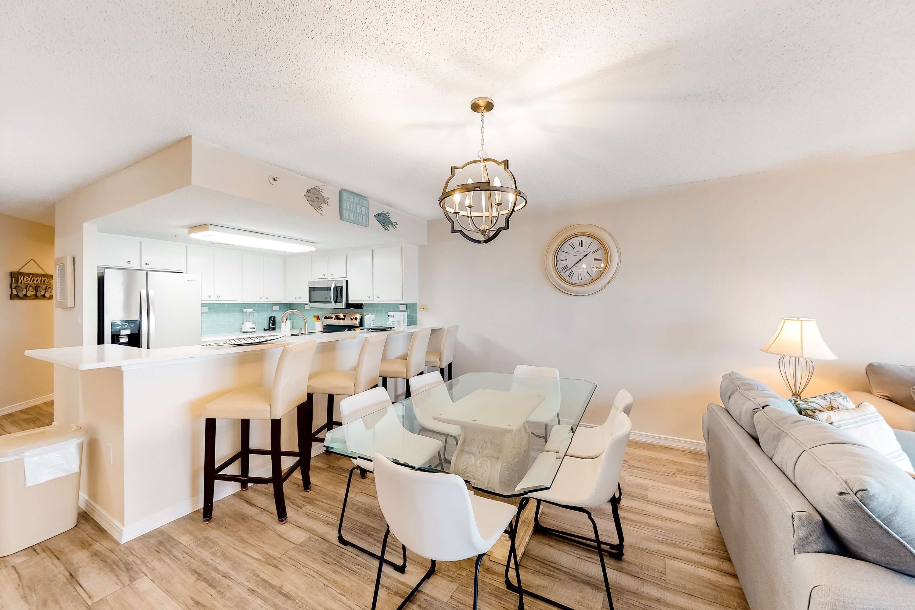 A modern kitchen with white cabinets and a central island, a dining area with a glass table and white chairs, and a living area with a sofa and lamp.
