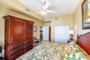A bedroom with a bed, a wooden wardrobe, a ceiling fan, and a door leading to another room.