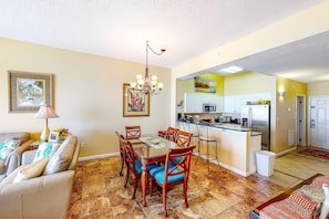 A dining area with a table and chairs, a chandelier, and a kitchen in the background.