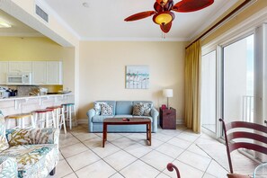 A living room with a sofa, coffee table, and a dining area with stools. There is a kitchen area with white cabinets and a microwave. A ceiling fan is visible, and the room has a large window with curtains.