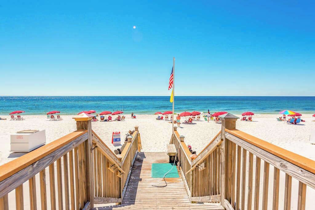 A wooden boardwalk leading to a beach with umbrellas and a flag.