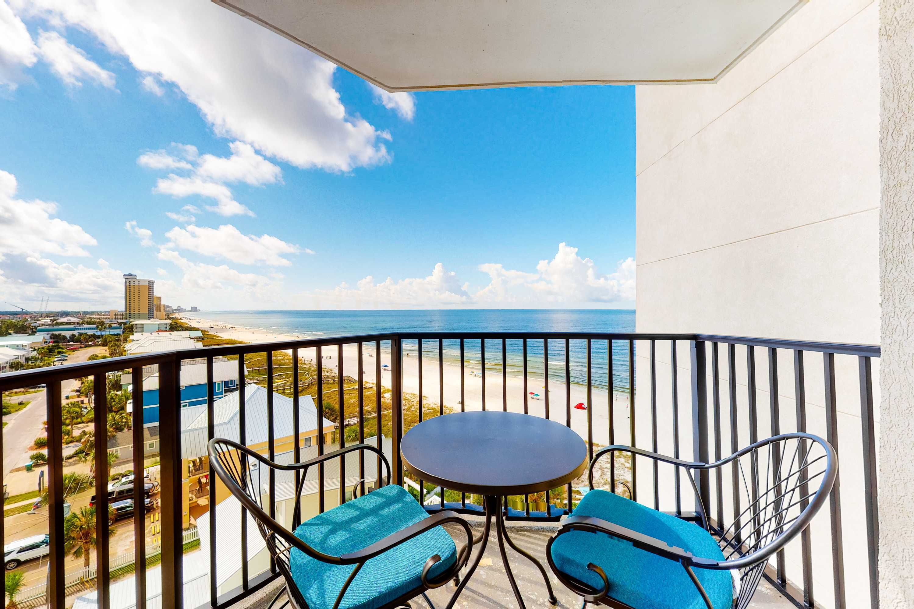 A balcony with a table and two chairs, overlooking a beach and ocean.