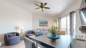 A modern living room with a dark wooden dining table, grey sofas, a ceiling fan, and a large window with curtains. - Condo with Pool Amenity in Miramar Beach (Miramar Beach)
