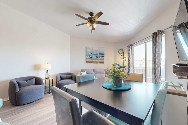 Een moderne woonkamer met een donker houten eettafel, grijze banken, een plafondventilator en een groot raam met gordijnen.
