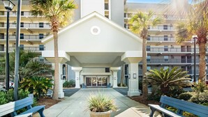 A building entrance with a covered walkway, benches, and palm trees. - Cozy beachfront condo with beach setup - near shopping (Fort Walton Beach)