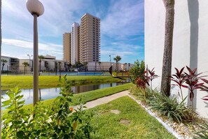 A view of a hotel with a tennis court, a small pond, and a tall building in the background. - Charming, dog-friendly condo with a pool, tennis, beach access, & washer/dryer (Panama City Beach)