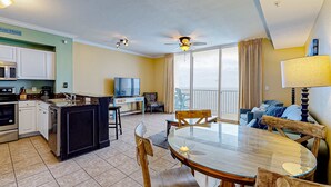 A modern kitchen with white cabinets, a microwave, and a dishwasher. There is a dining area with a glass table and wooden chairs. A ceiling fan and a TV are visible in the room. - 21st-floor oceanfront condo with ocean view, pool & hot tub access & W/D (Panama City Beach)