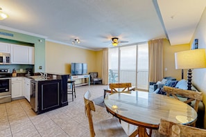 A modern kitchen with white cabinets, a microwave, and a dishwasher. There is a dining area with a glass table and wooden chairs. A ceiling fan and a TV are visible in the room. - 21st-floor oceanfront condo with ocean view, pool & hot tub access & W/D (Panama City Beach)