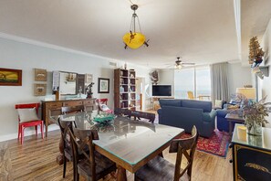 A spacious living room with a glass-top dining table, a blue sofa, and a bookshelf. - Spacious condo with full kitchen, shared pool and beachfront (Panama City Beach)