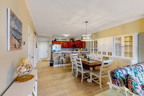A spacious kitchen with wooden cabinets, a dining area with white chairs, and a large window with a view. - Snowbird friendly gulf-view condo w/ Pool  Hot tub, & Private Balcony (Panama City Beach)