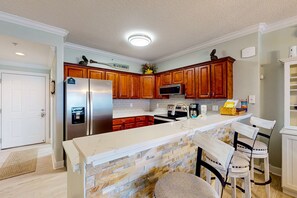 A kitchen with wooden cabinets, a granite countertop, a refrigerator, and a microwave. - Snowbird friendly gulf-view condo w/ Pool  Hot tub, & Private Balcony (Panama City Beach)