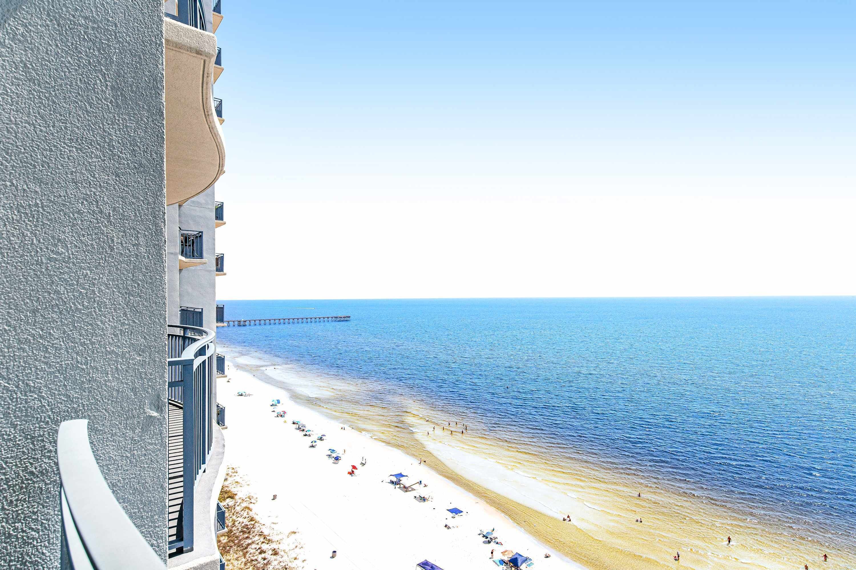A beach view with a pier extending into the sea, a sandy beach with umbrellas, and a building with balconies overlooking the beach.