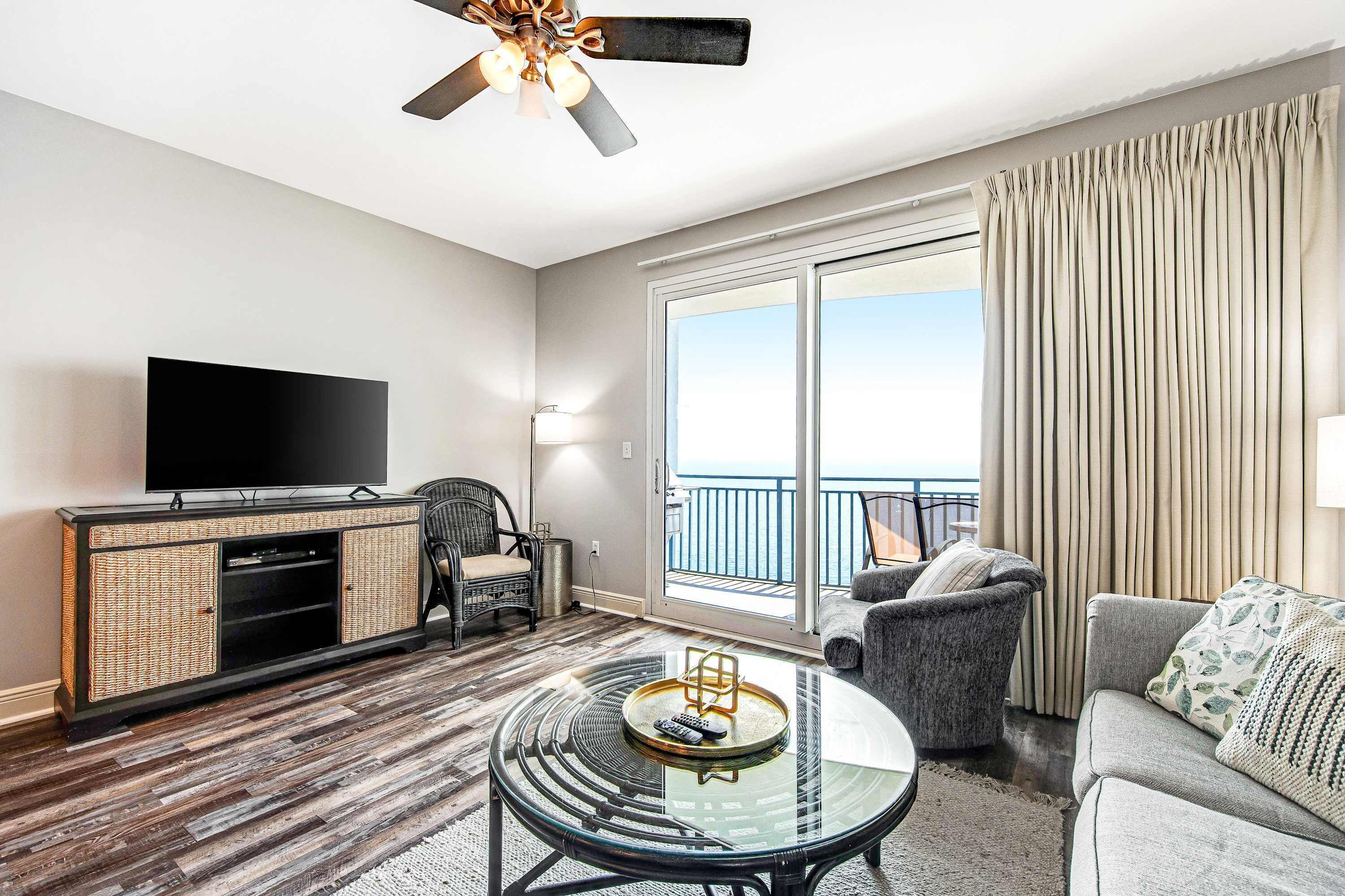 A modern hotel room with a flat-screen TV, wicker furniture, a glass coffee table, and a balcony with curtains.
