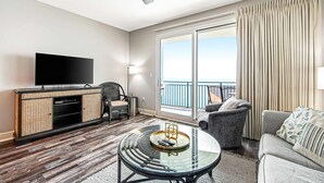 Een moderne hotelkamer met een flatscreen-televisie, rieten meubels, een glazen salontafel en een balkon met gordijnen.