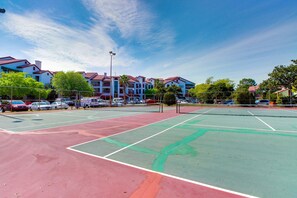 A tennis court with a net, surrounded by a parking area and buildings with red roofs.