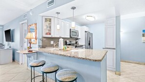 A modern kitchen with granite countertops, white cabinets, a microwave, and a refrigerator. There are three bar stools with patterned cushions. - Comfortable beachfront condo with beach service - quick drive to dining (Fort Walton Beach)