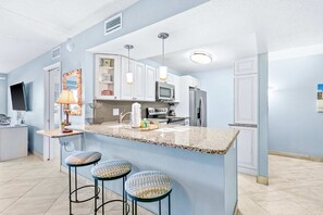 A modern kitchen with granite countertops, white cabinets, a microwave, and a refrigerator. There are three bar stools with patterned cushions. - Comfortable beachfront condo with beach service - quick drive to dining (Fort Walton Beach)