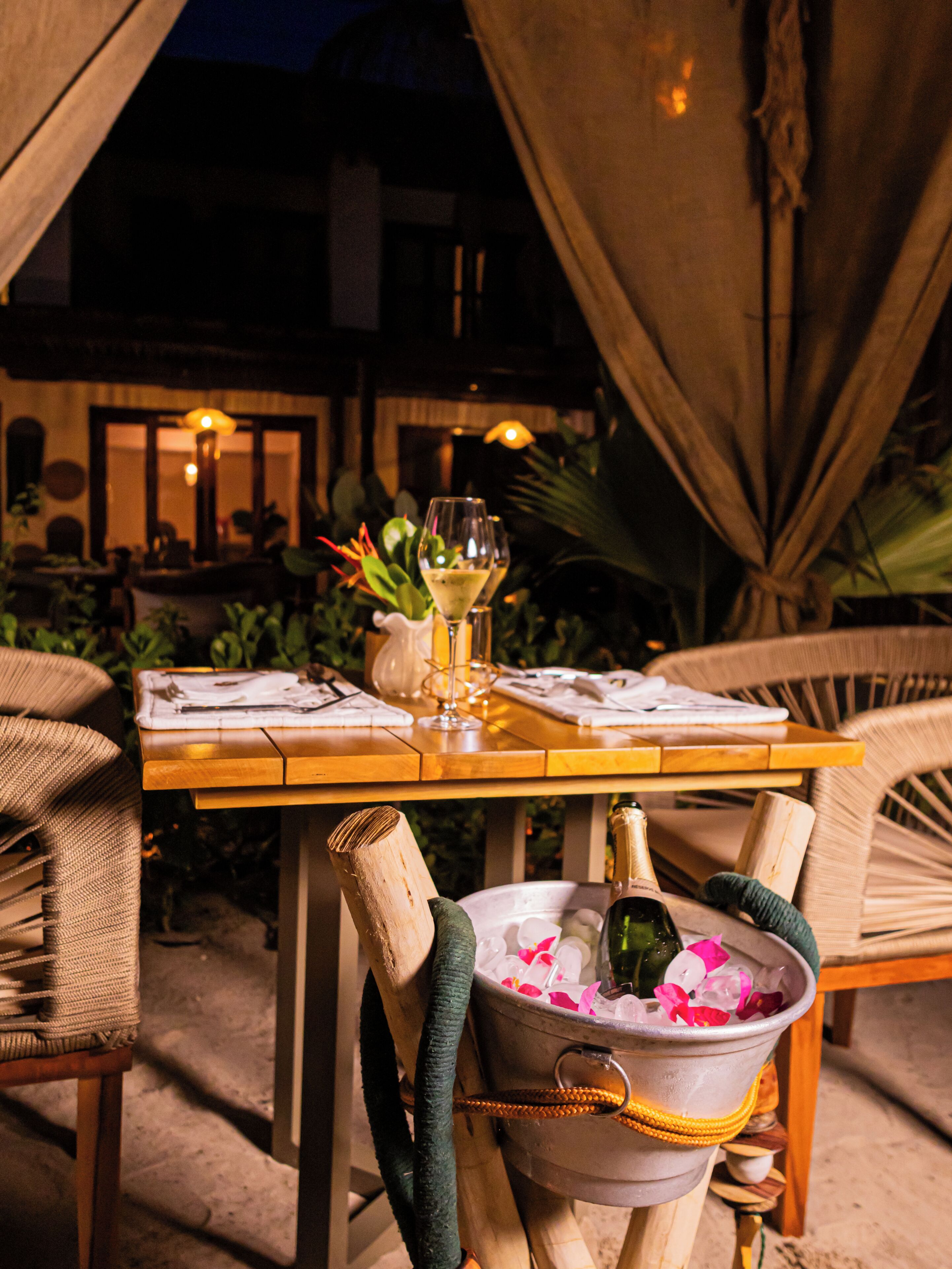 A dining table set outdoors with a bucket of ice and a bottle of champagne.