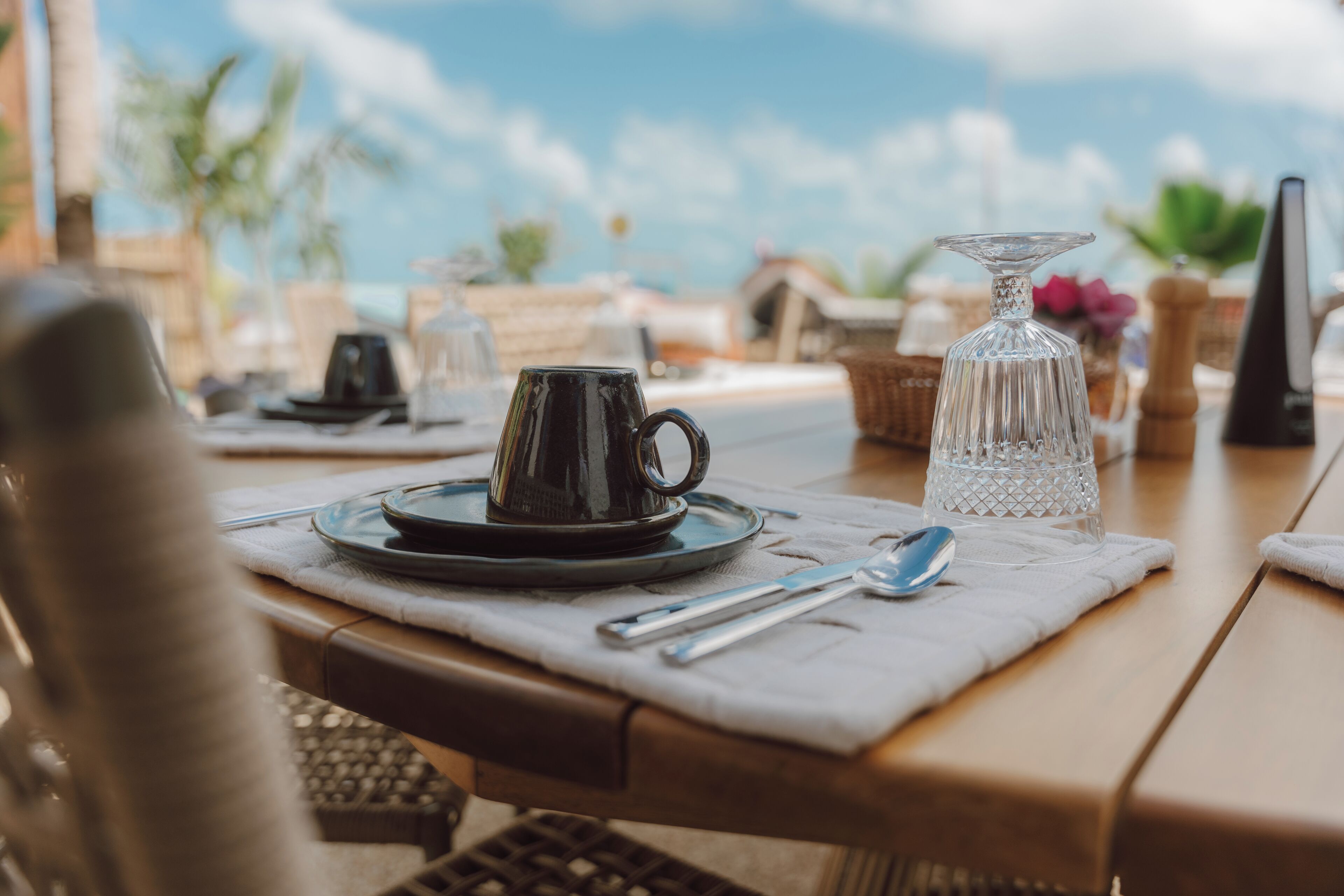 Une table dressée avec une tasse à café, un verre et des couverts sur une nappe, offrant une vue dégagée sur le ciel et quelques éléments de végétation en arrière-plan.