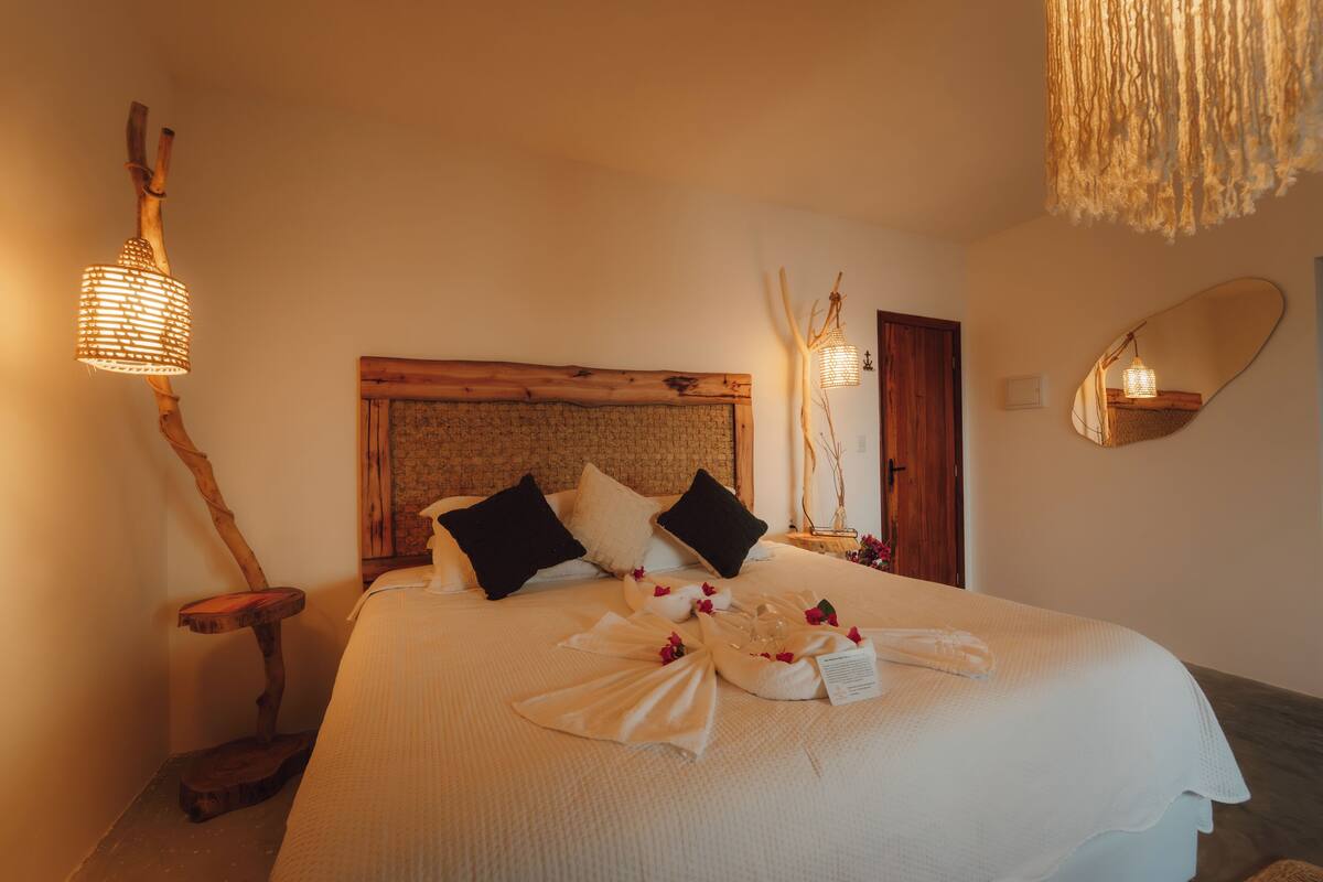 Een opgemaakt bed met een houten hoofdbord, twee zwarte kussens en een witte deken. Twee wandlampen met een gevlochten ontwerp, een houten nachtkastje en een spiegel aan de muur.