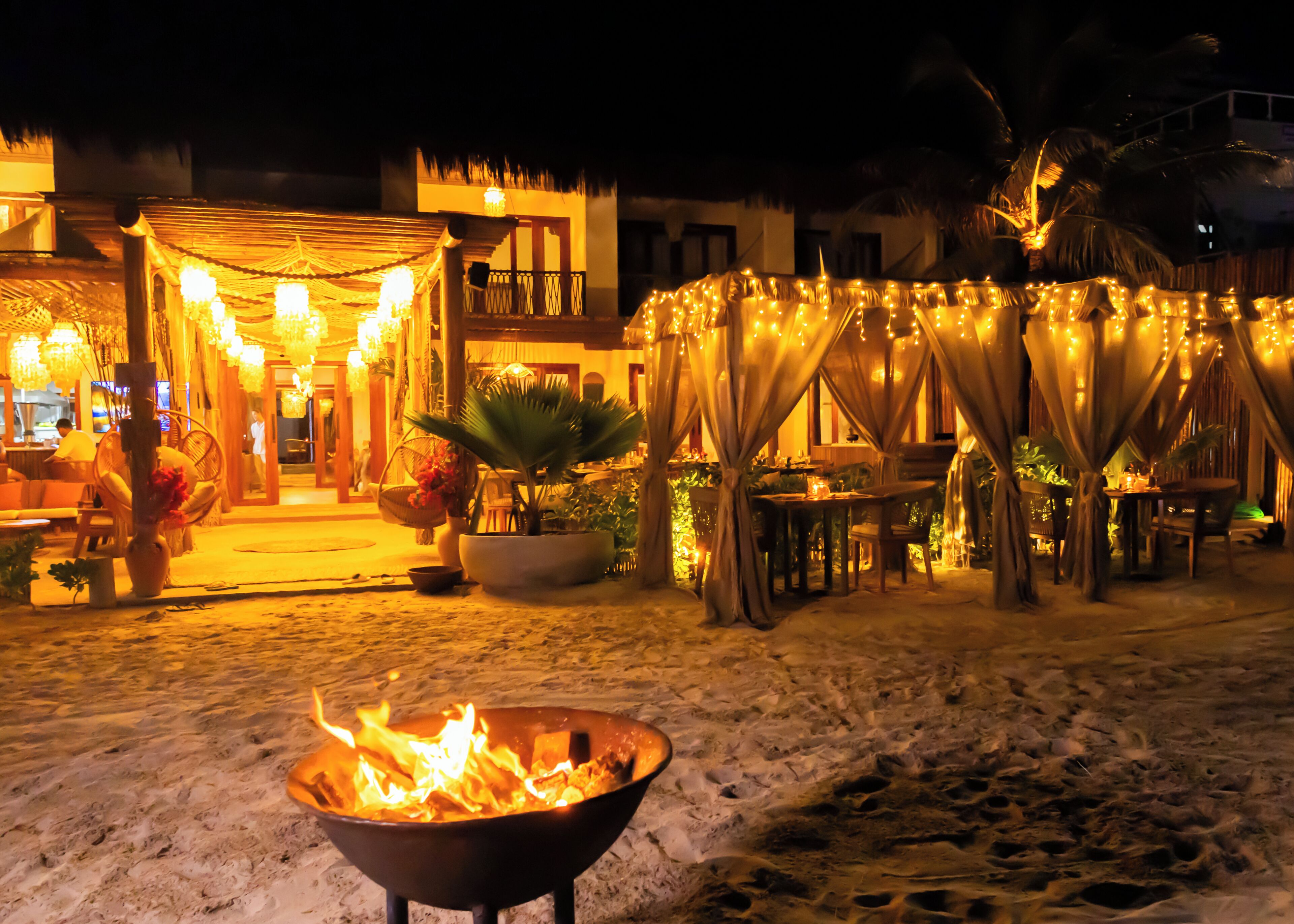 A nighttime outdoor area with a fire pit, tables, chairs, and string lights.