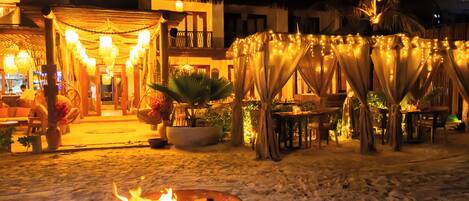 A nighttime outdoor area with a fire pit, tables, chairs, and string lights.