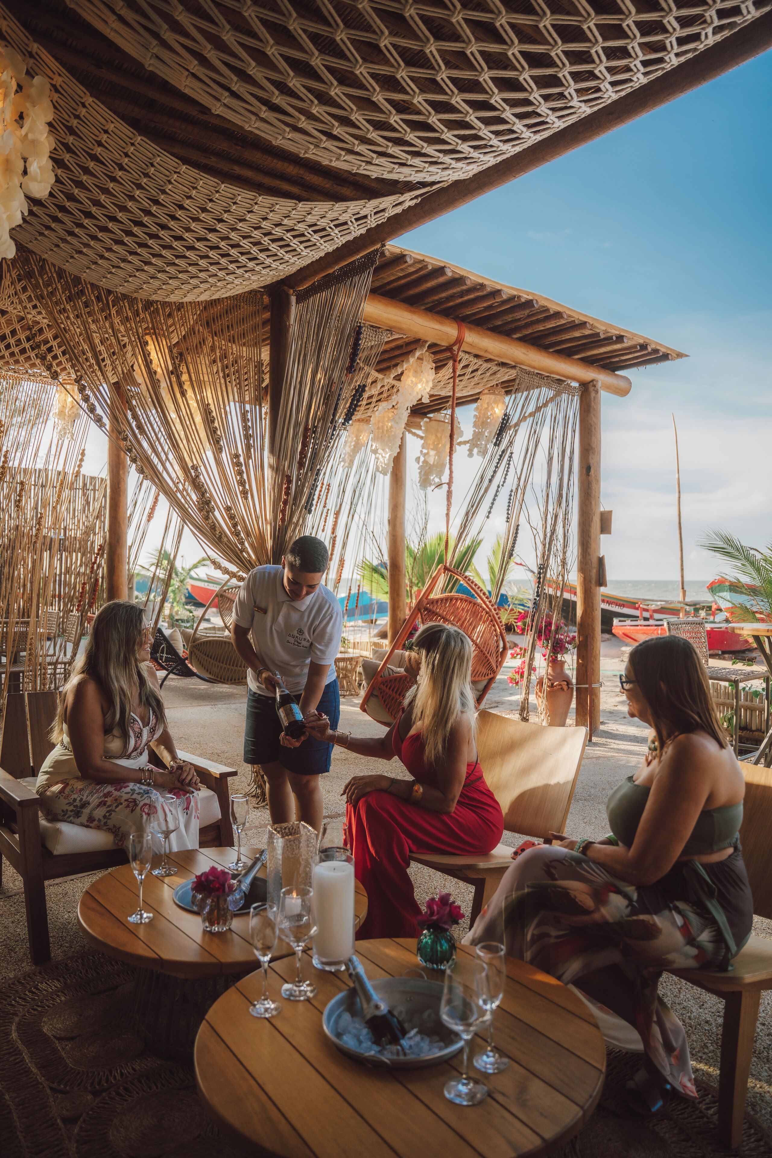 A group of people seated at a wooden table with drinks, under a woven canopy.