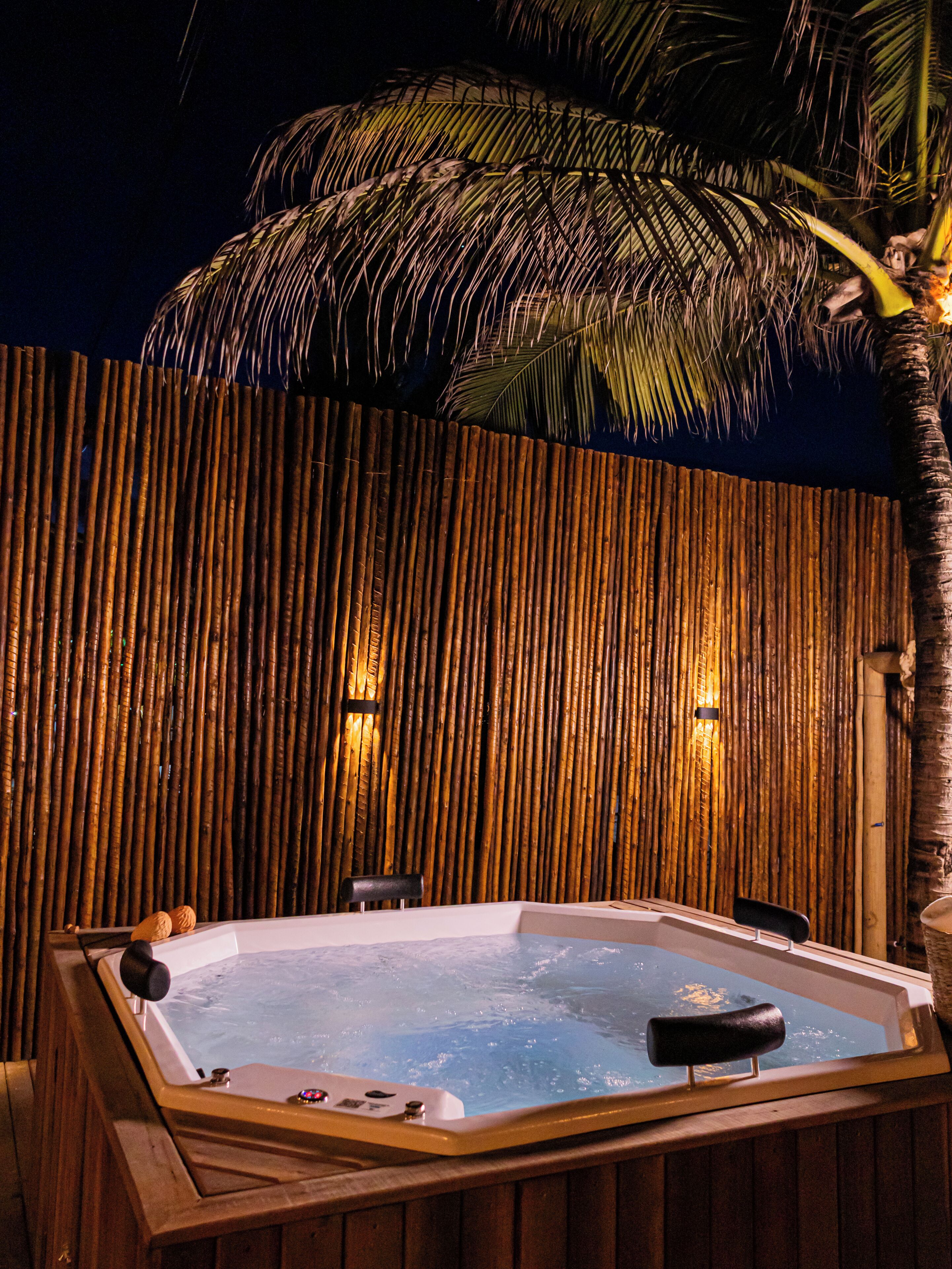 A hot tub with a wooden deck, a bamboo wall, and a palm tree.