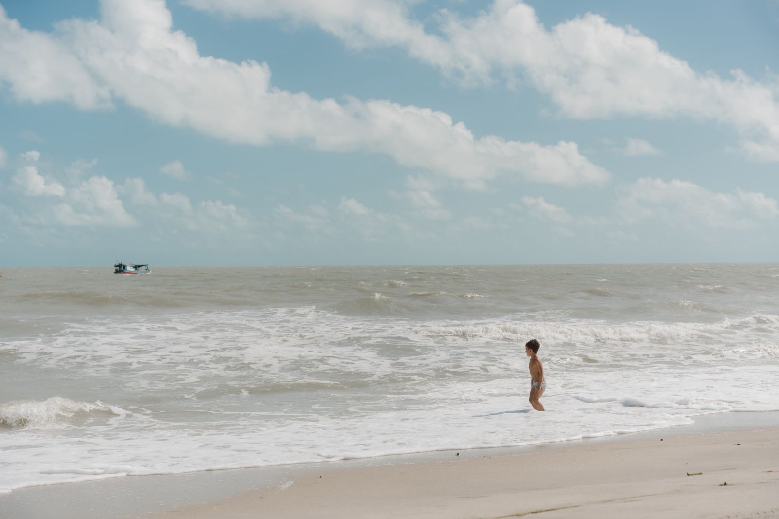 A beach scene with a child standing in the water, waves, and a distant boat.